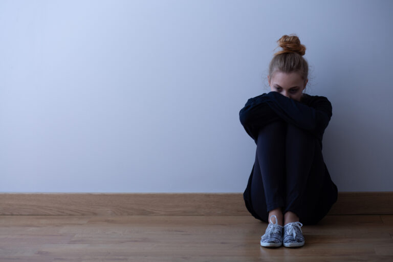 Young woman with anxiety disorder wearing dark clothes sitting on the floor, copy space on empty wall