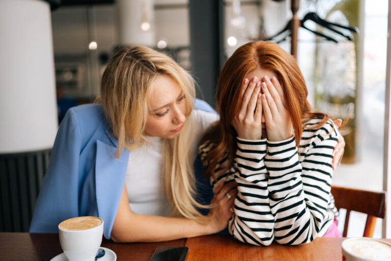 Portrait of displeased young woman and best female friend trying to comfort and cheer up sitting together in cafe by window